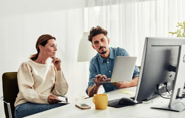 Business settings. Two colleagues seated side by side by a desk. The man holds a laptop, showing his female colleague business content.